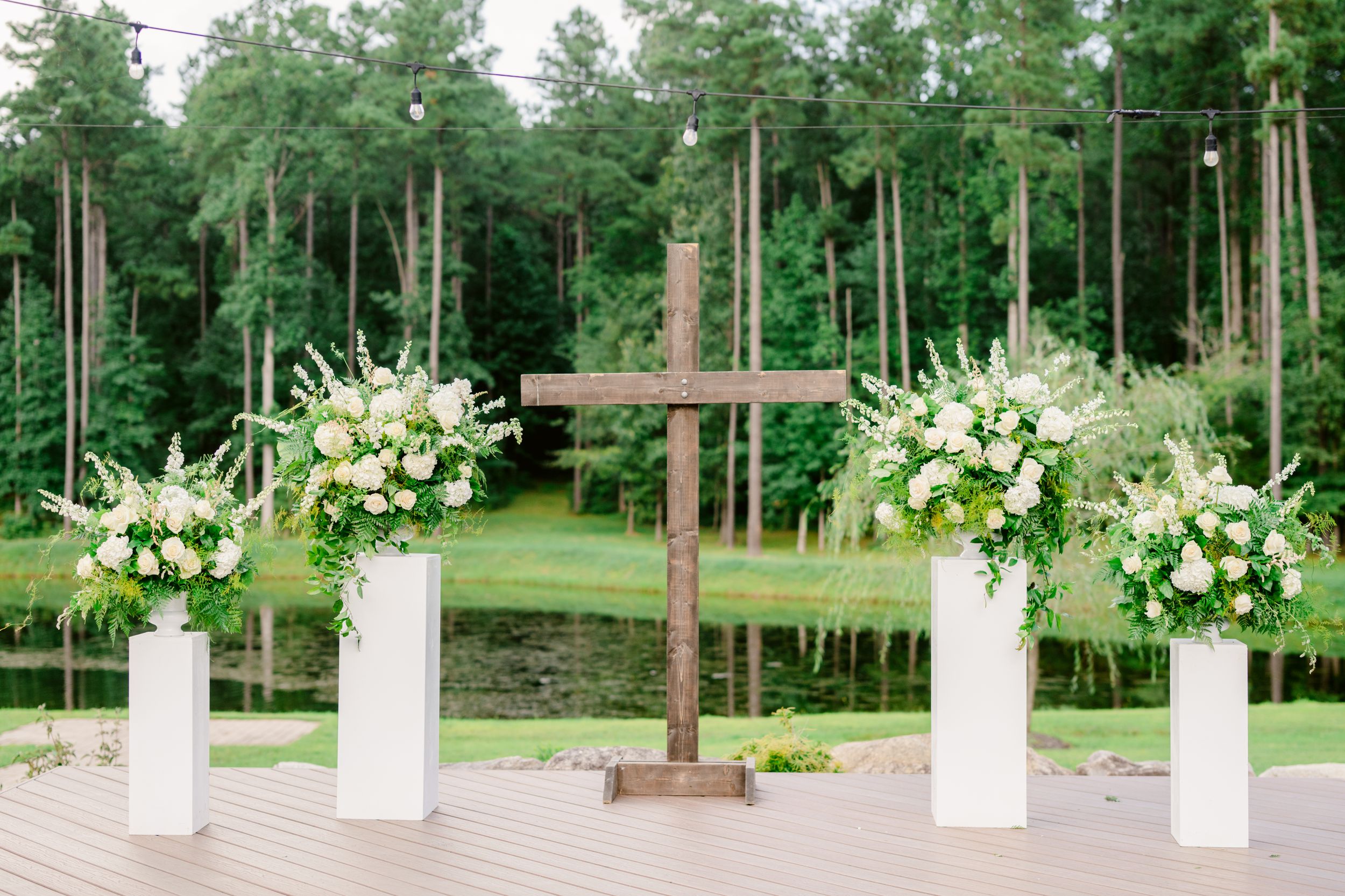 Wooden cross and white floral arrangements on pedestals create an elegant outdoor wedding ceremony setup in a pine forest.