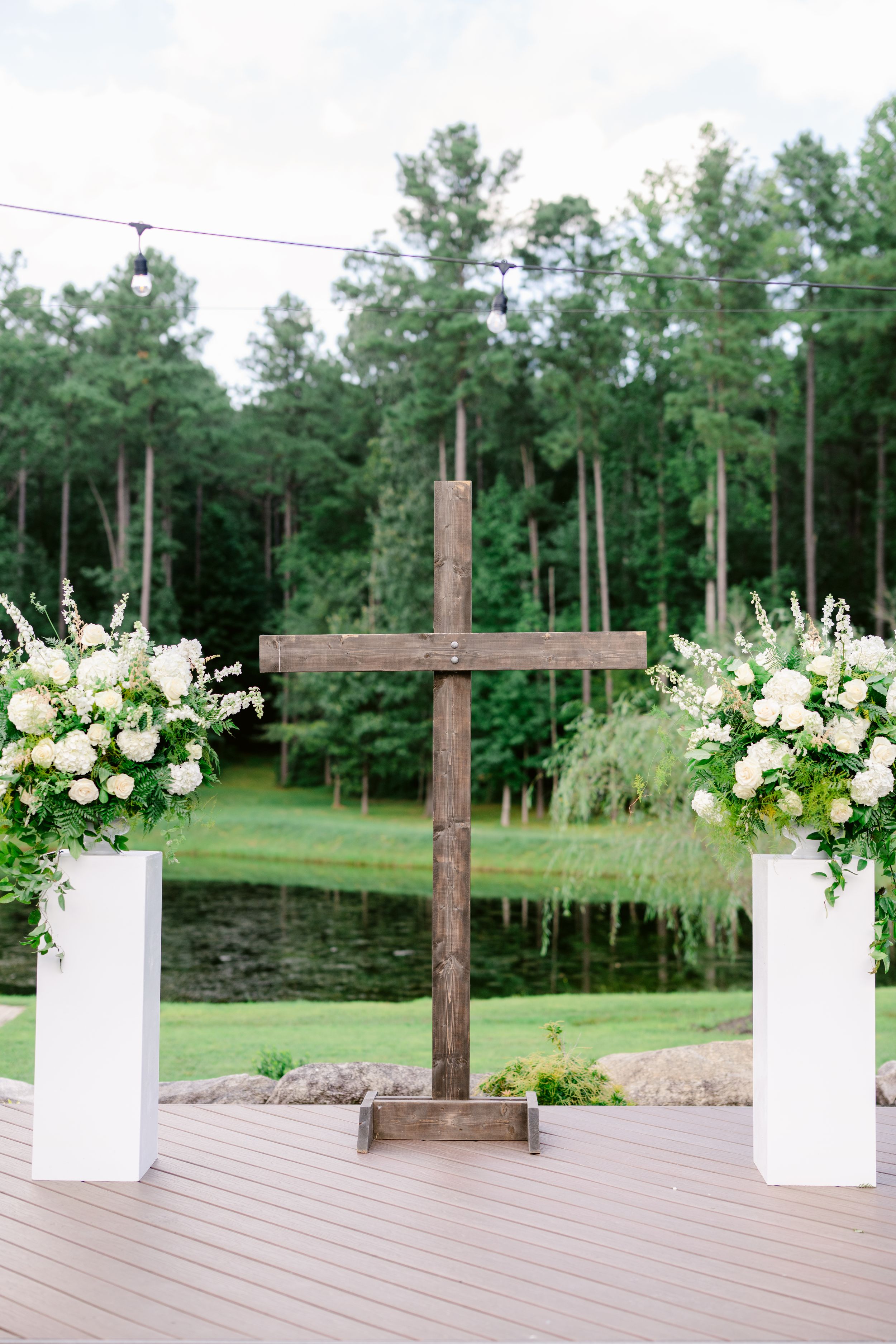 Wooden crosses and white floral arrangements line a rustic outdoor wedding ceremony space with pine trees in background.