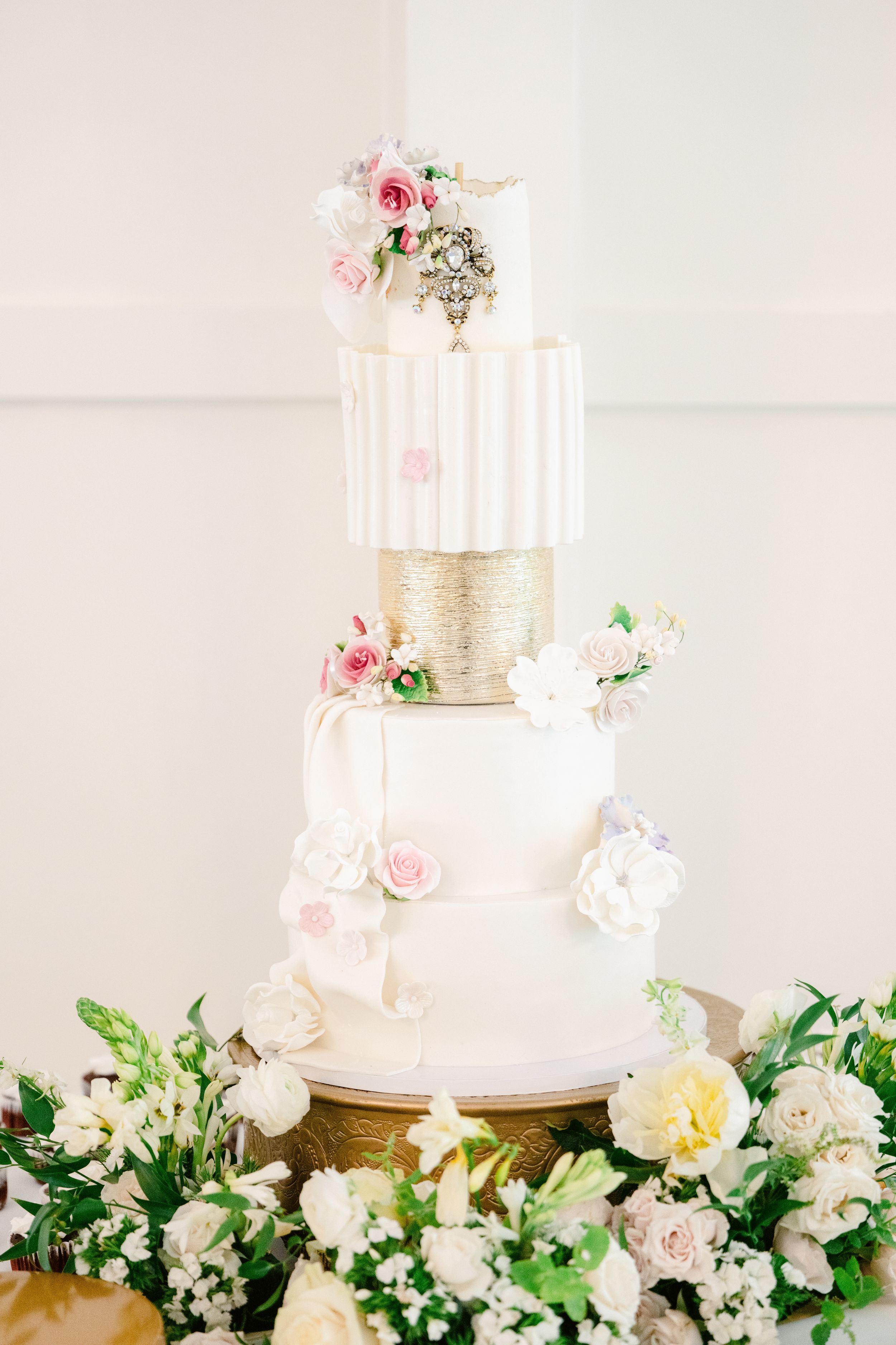 Elegant white wedding cake with pink roses and gold accents displayed on a floral-decorated dessert table.