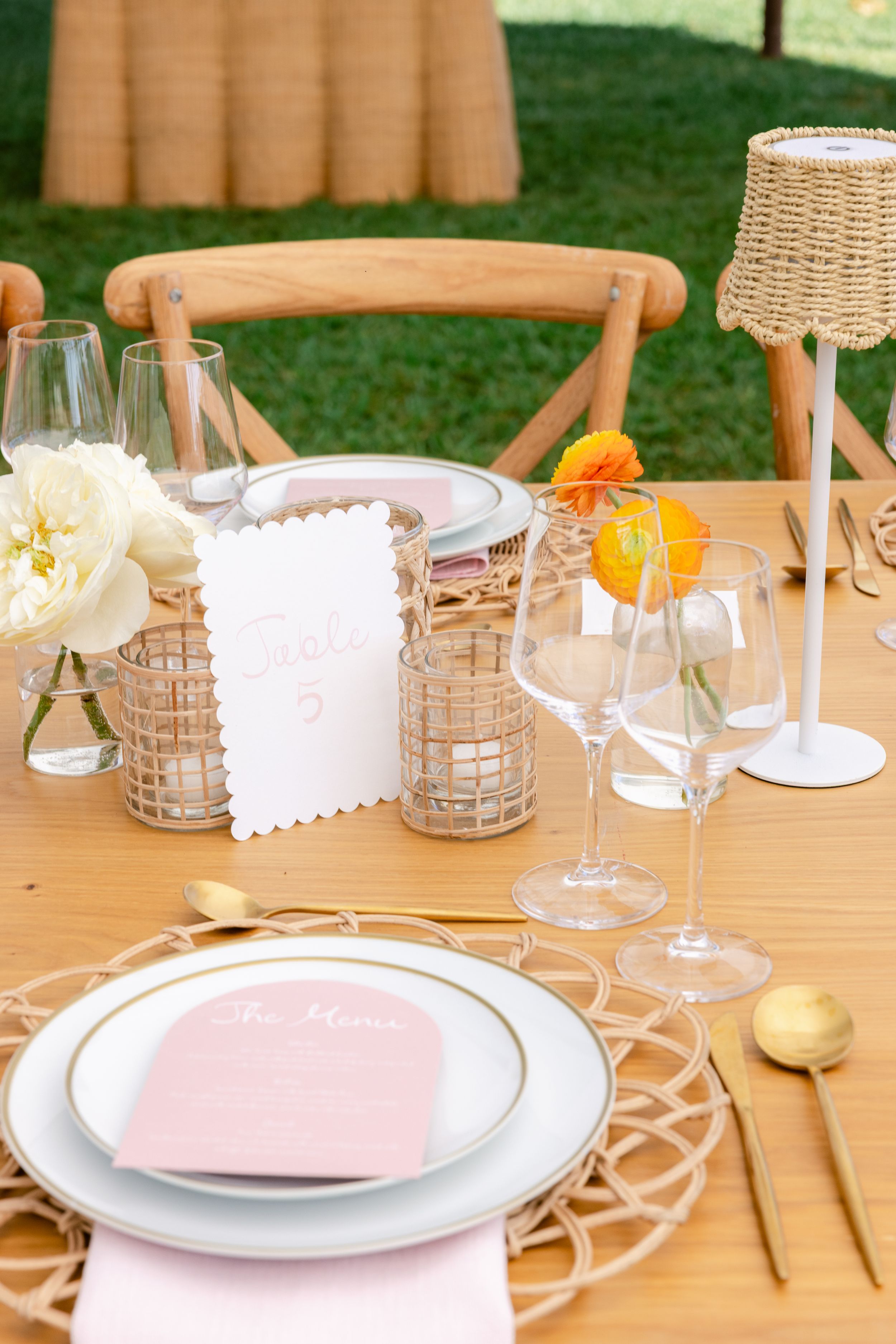 Elegant outdoor table setting with wooden chairs, white plates, pink napkins, and small floral centerpieces in glass vases.