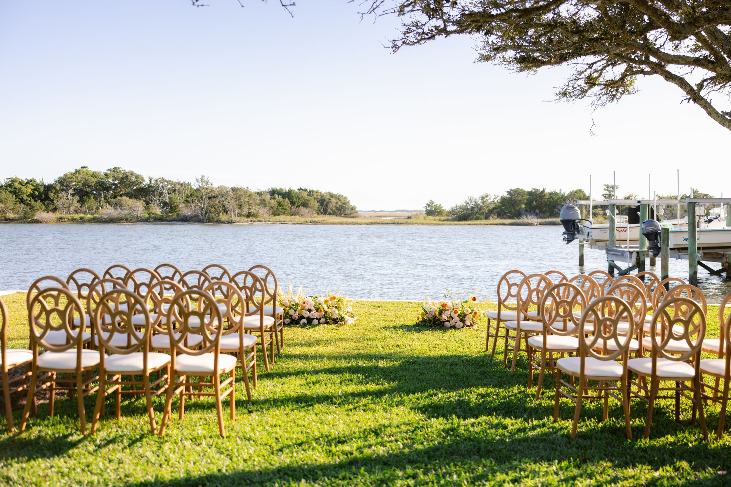 Wooden cross-back chairs arranged in rows on a grassy lawn overlooking a waterfront for an outdoor wedding ceremony.