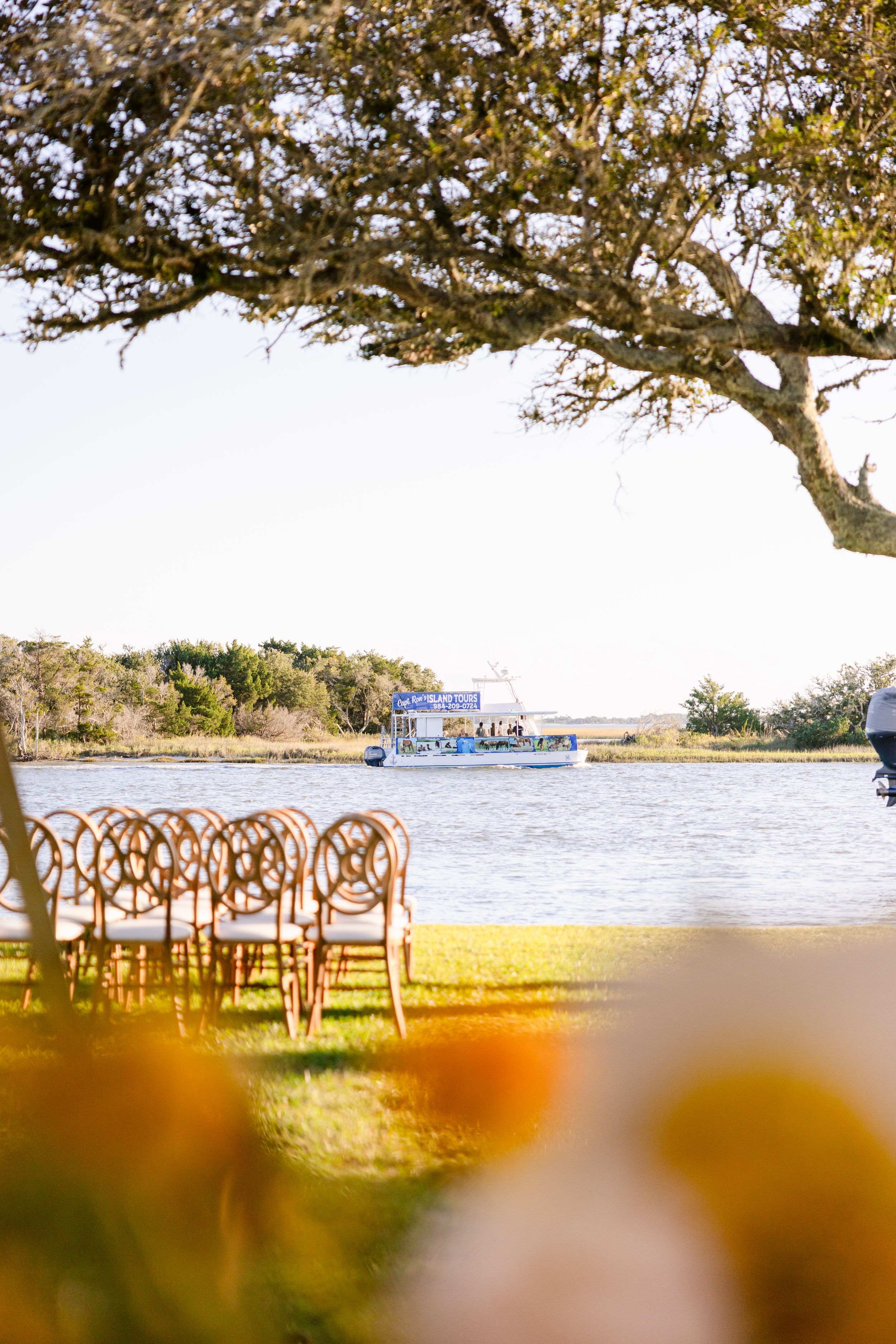 Panoramic view of a waterfront wedding venue with wooden chairs arranged under a tree by the ocean.
