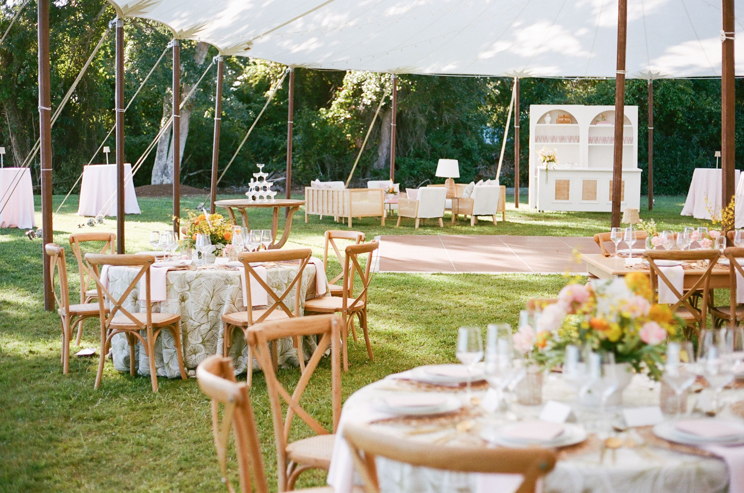 Elegant outdoor wedding reception setup with wooden cross-back chairs and white linens under string lights at sunset.