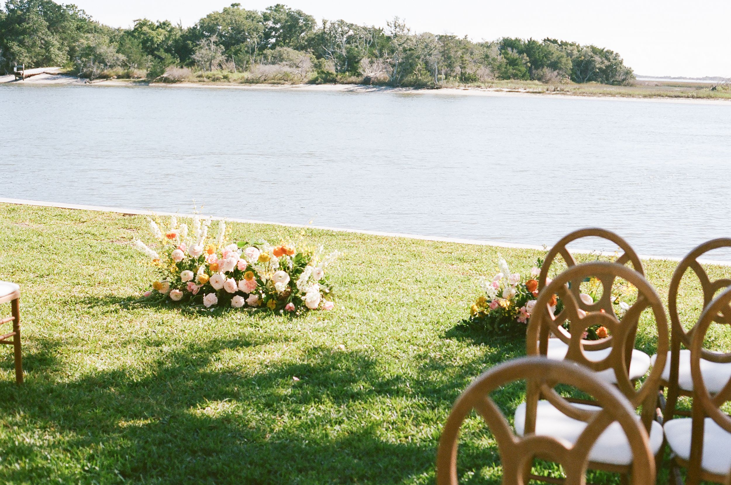 Wooden cross-back chairs arranged in rows on grass with floral decorations for an outdoor ceremony.