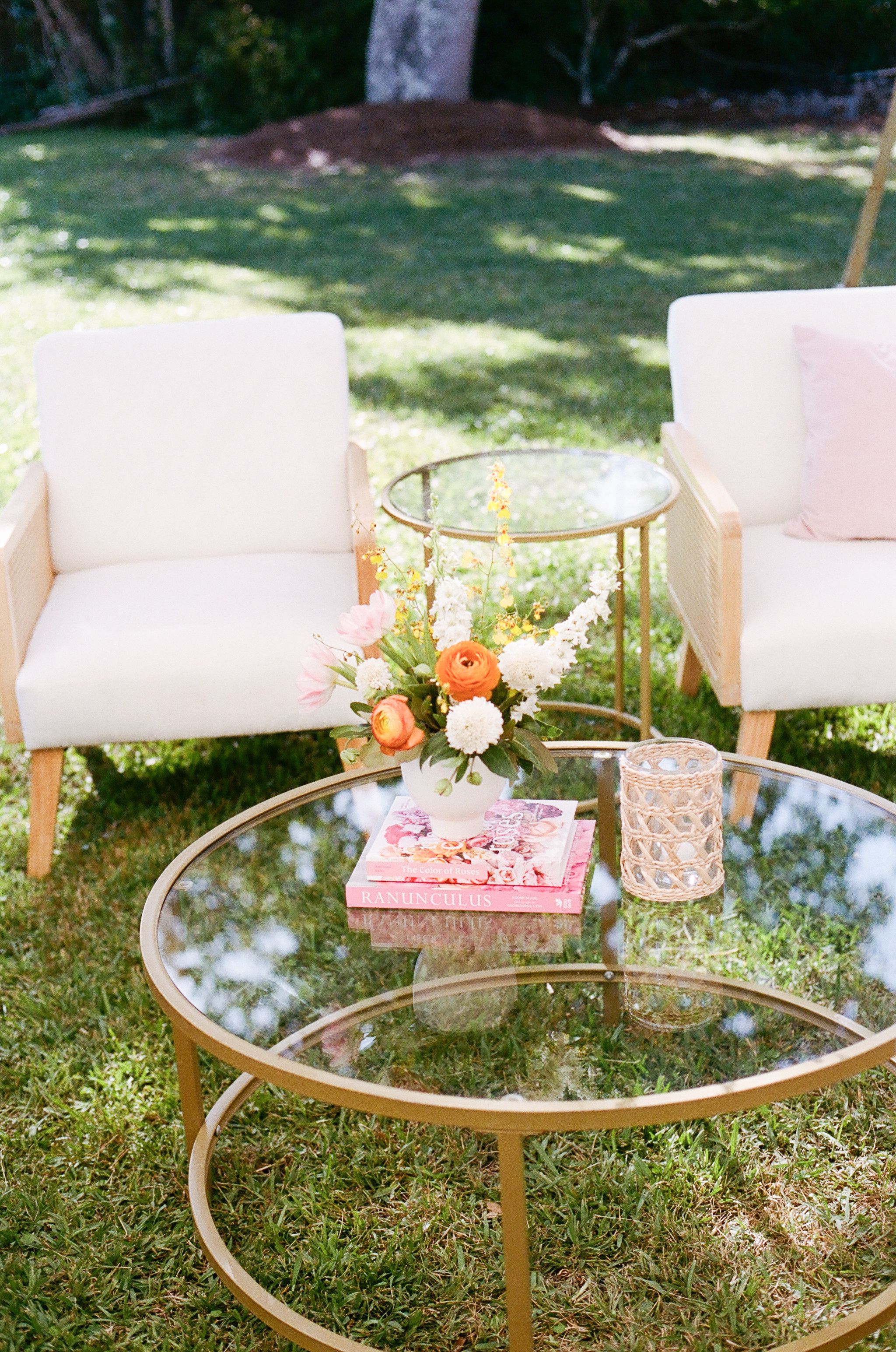 Round glass coffee tables with gold frames arranged on grass with white cushioned seating for an outdoor gathering.