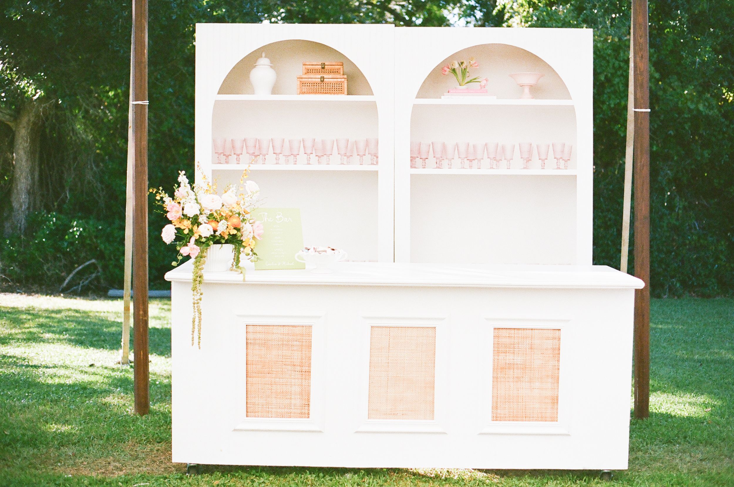 White vintage-style bar cabinet with arched shelving displays glassware at outdoor wedding.