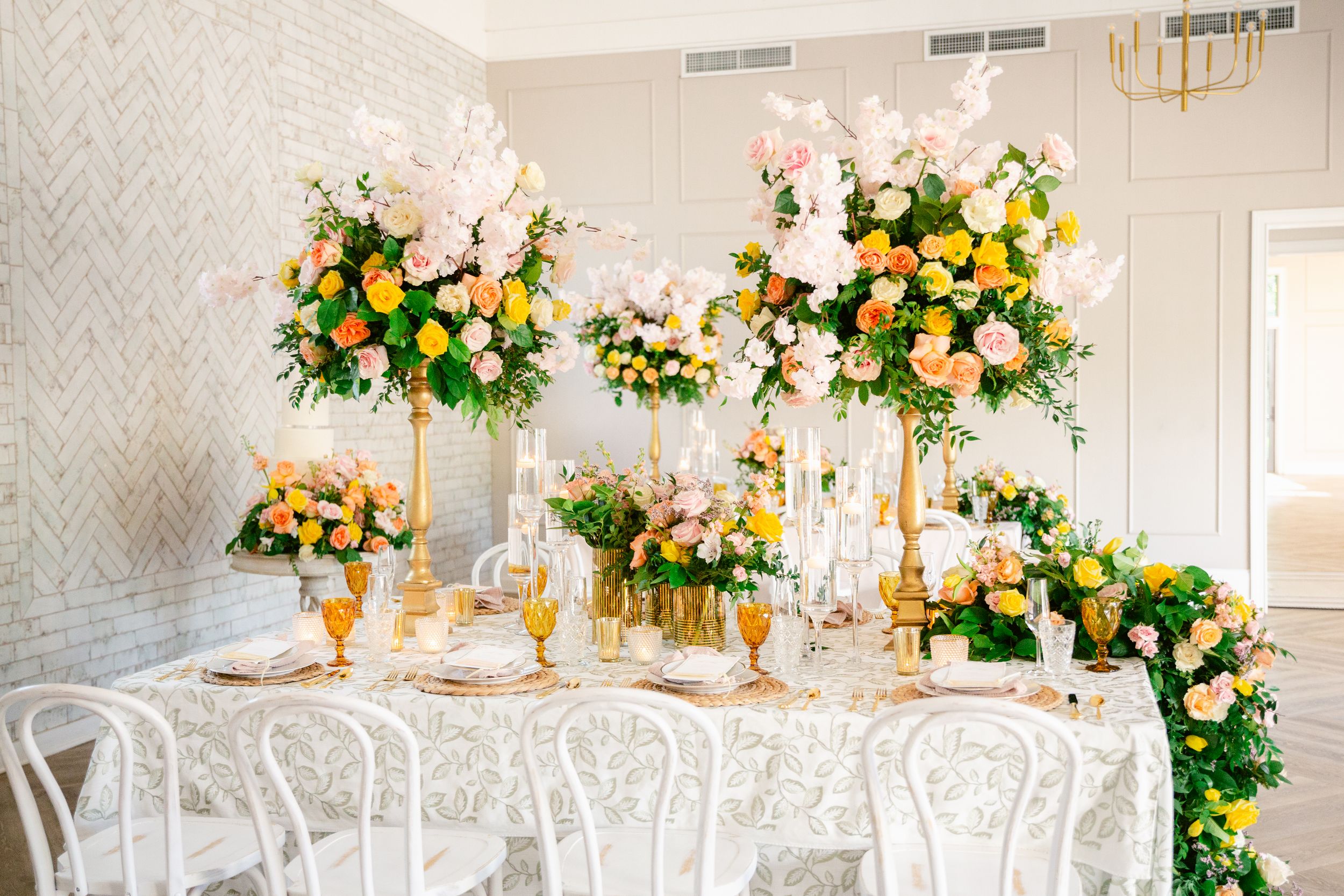 Elegant white wedding reception tables with floral centerpieces featuring pink and peach roses in a light-filled ballroom.