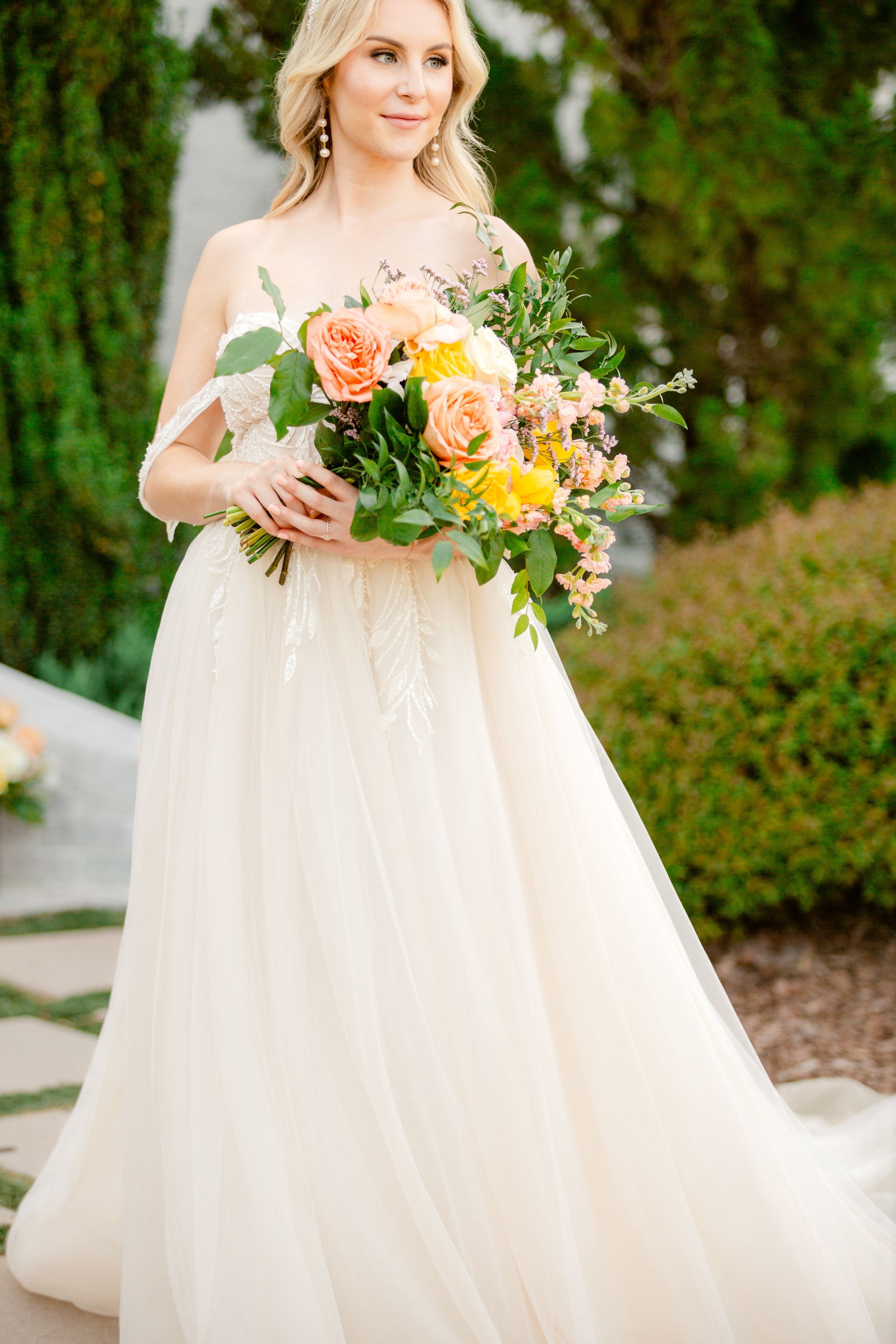 A bride in a flowing white gown holds a vibrant coral and yellow floral bouquet against a lush garden backdrop.