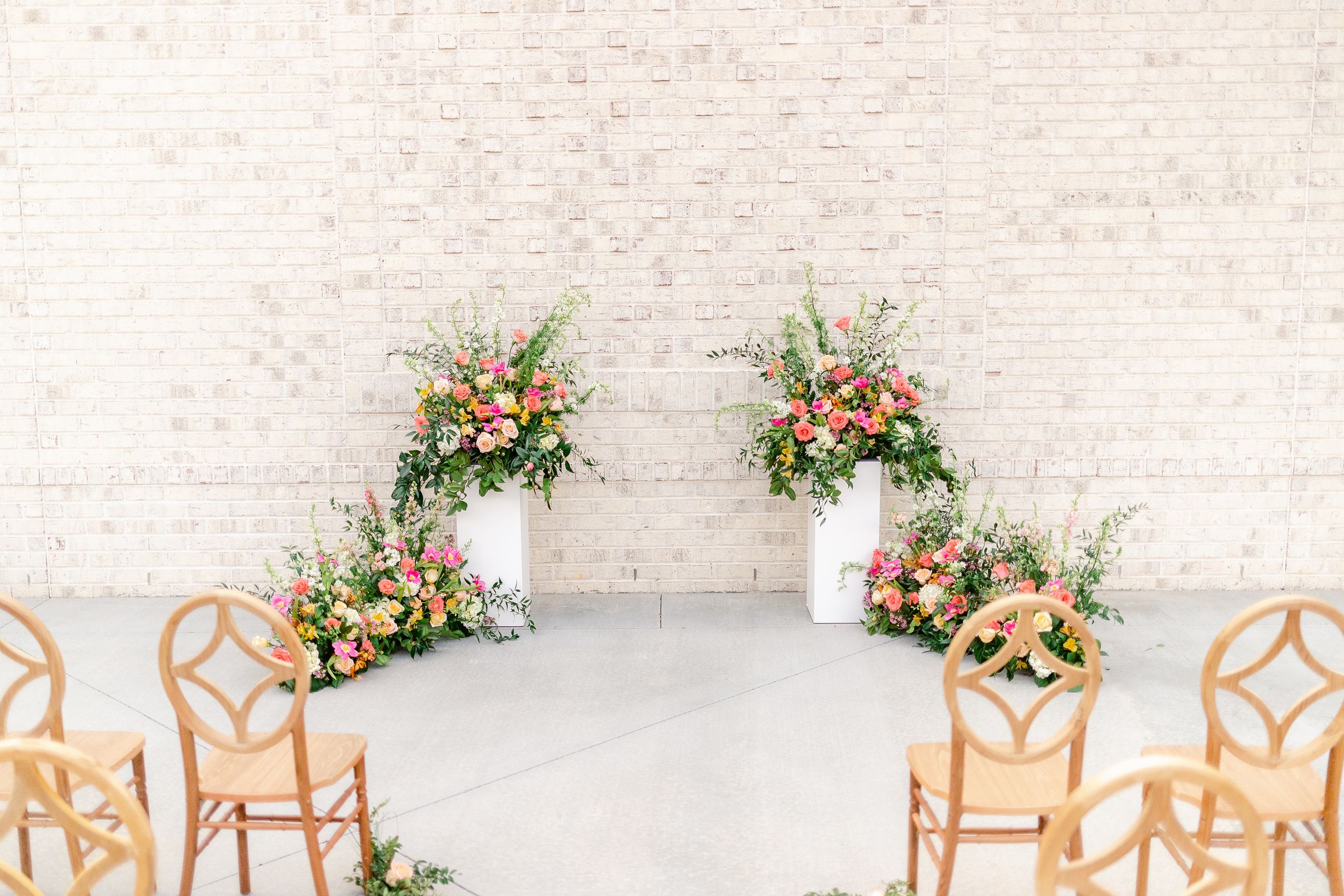 Elegant wedding ceremony setup with white chairs and floral arrangements against a textured stone wall.