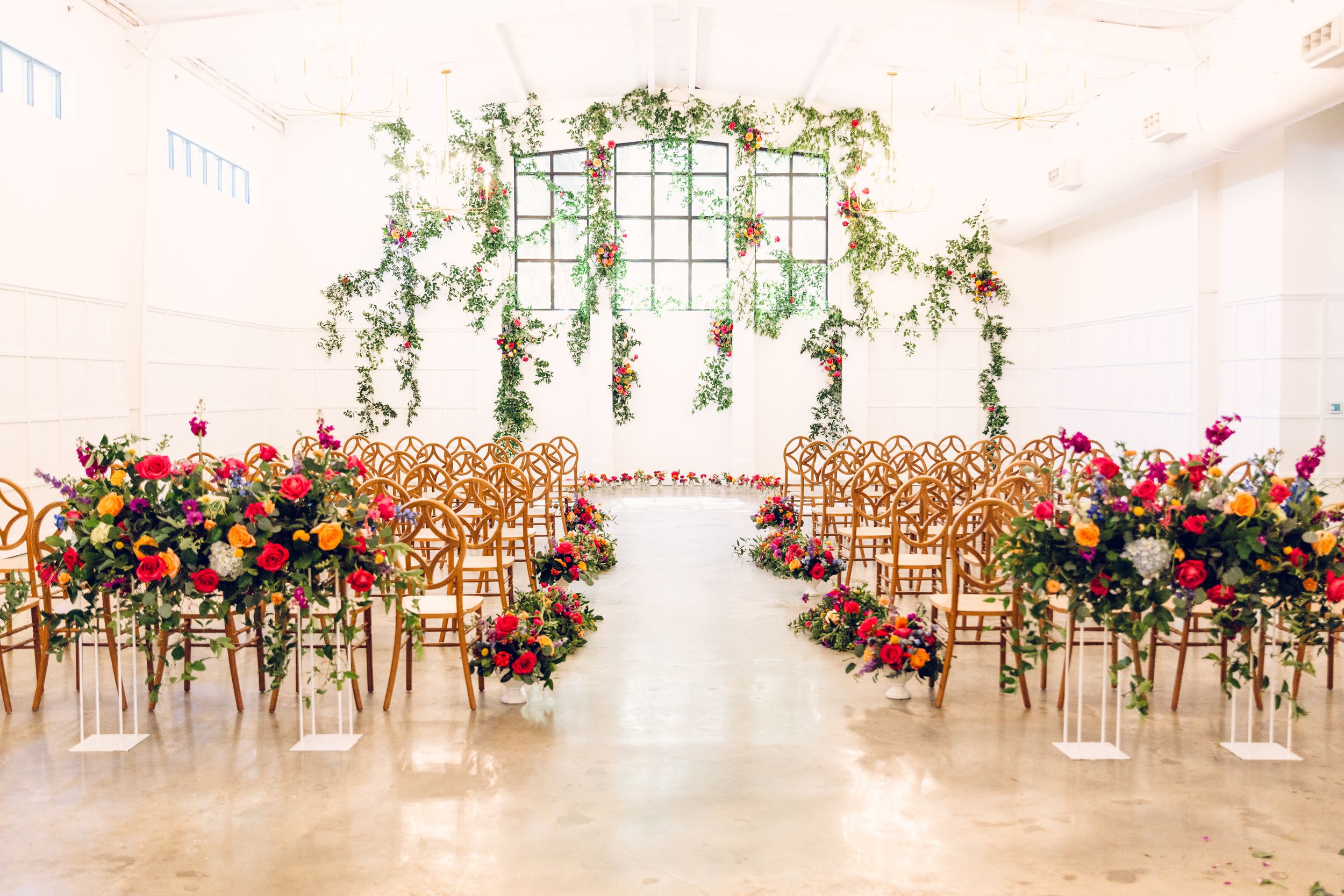 A wedding ceremony setup with wooden chairs, floral arrangements, and hanging greenery in a bright white venue.
