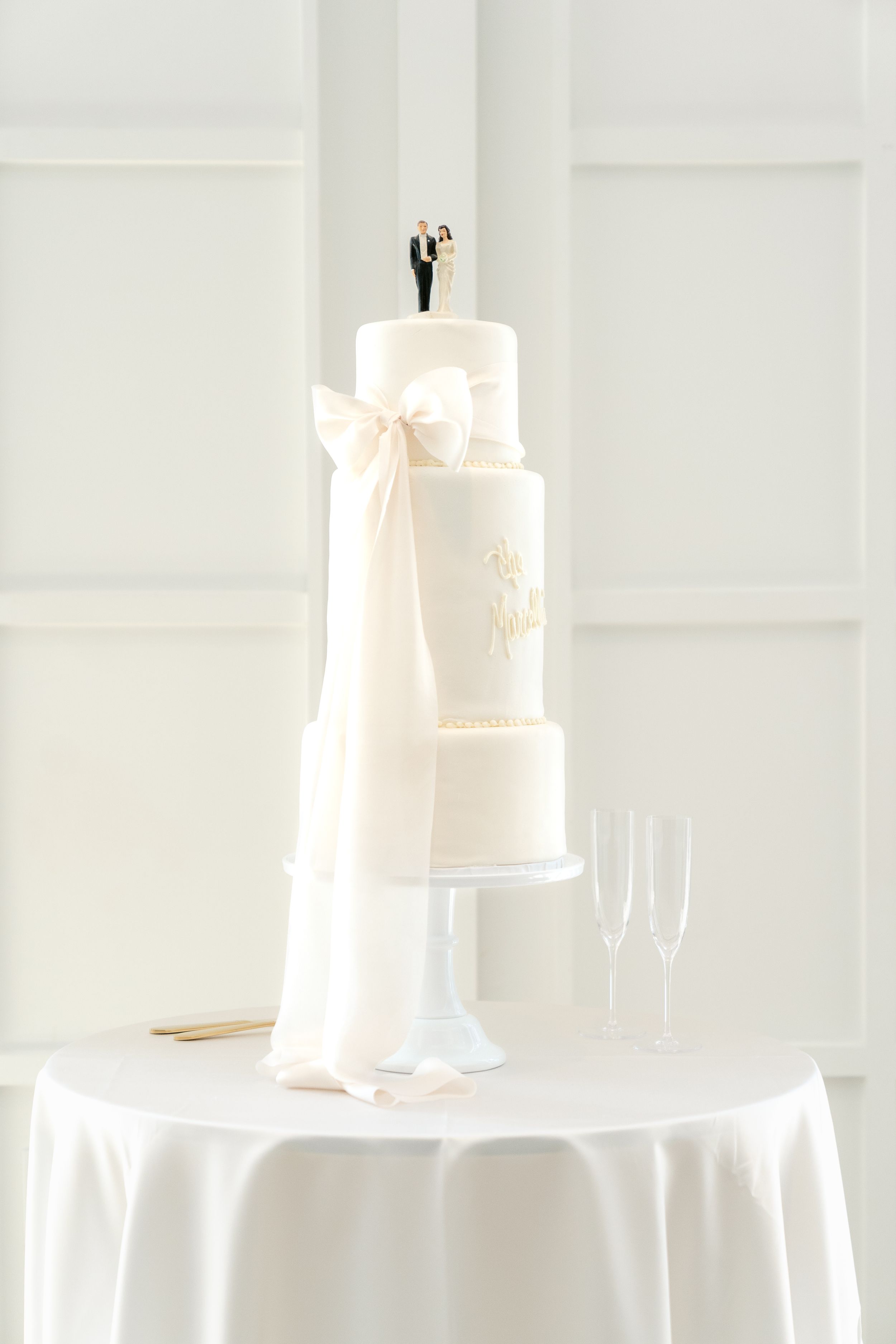 Elegant white wedding cake with multiple tiers displayed on white tablecloth with champagne glasses in background.