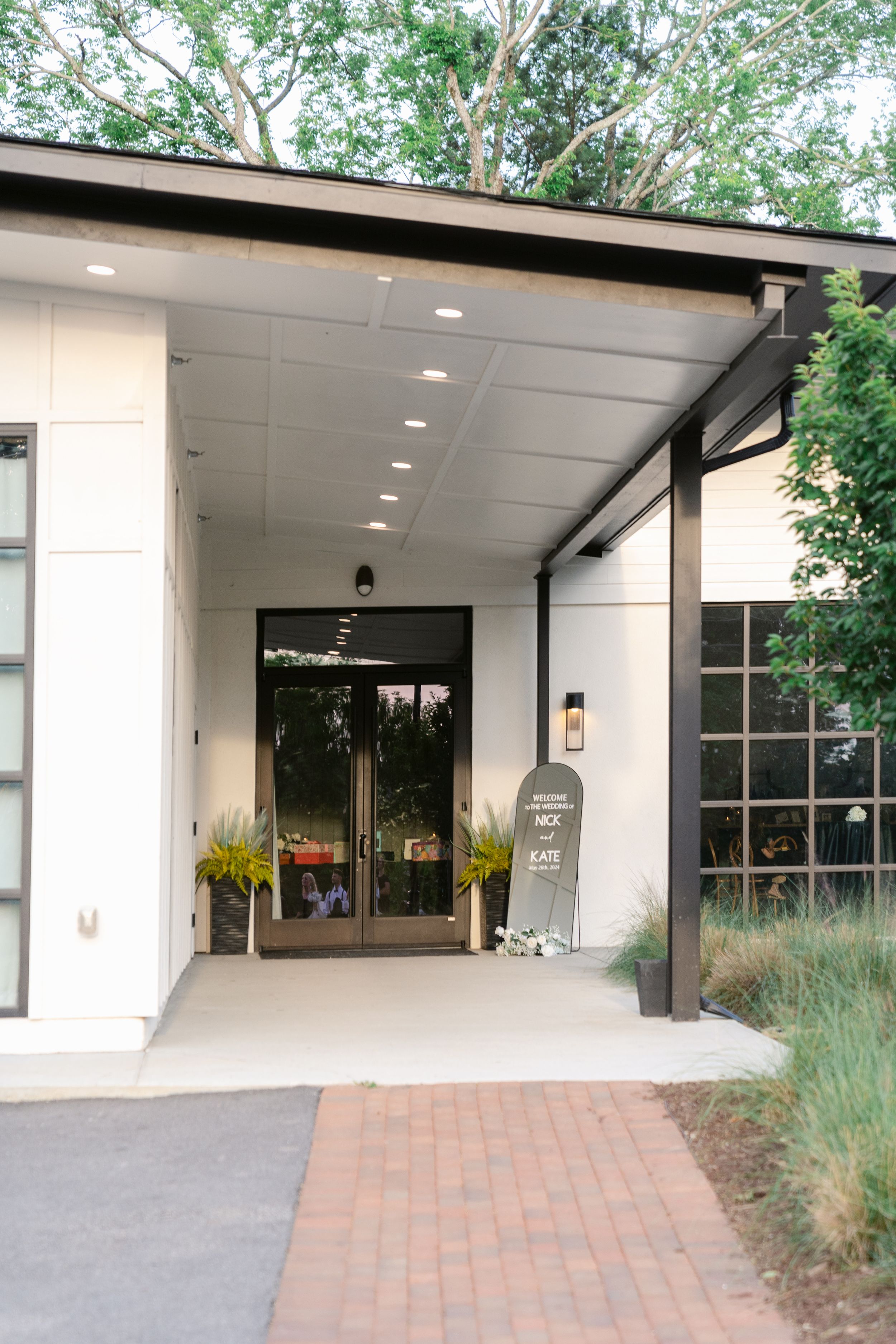Modern building entrance with covered walkway, white walls, recessed lighting and brick paved path leading to glass doors.