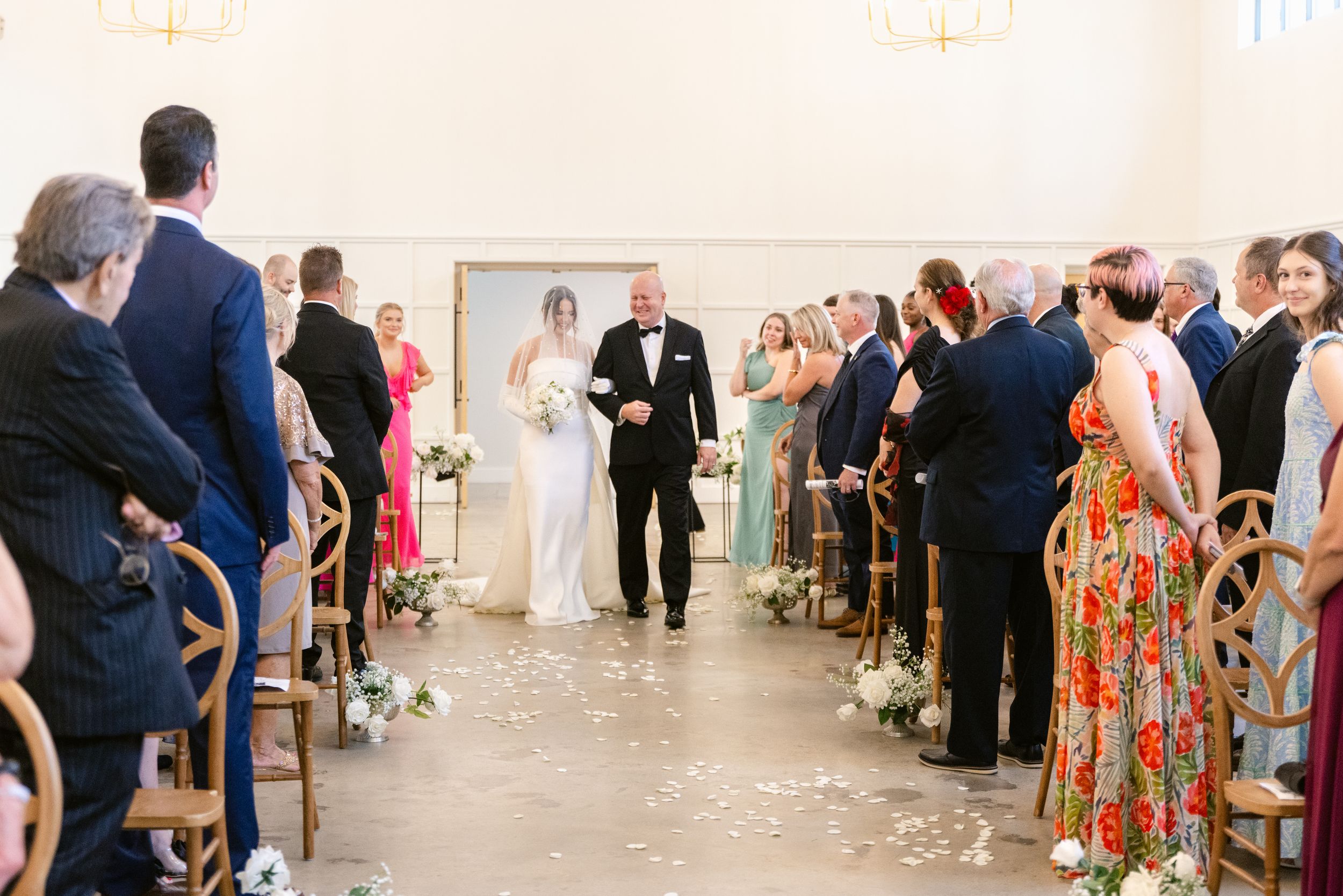 Wedding guests stand and watch as a couple walks down the aisle in a bright white ceremony space.