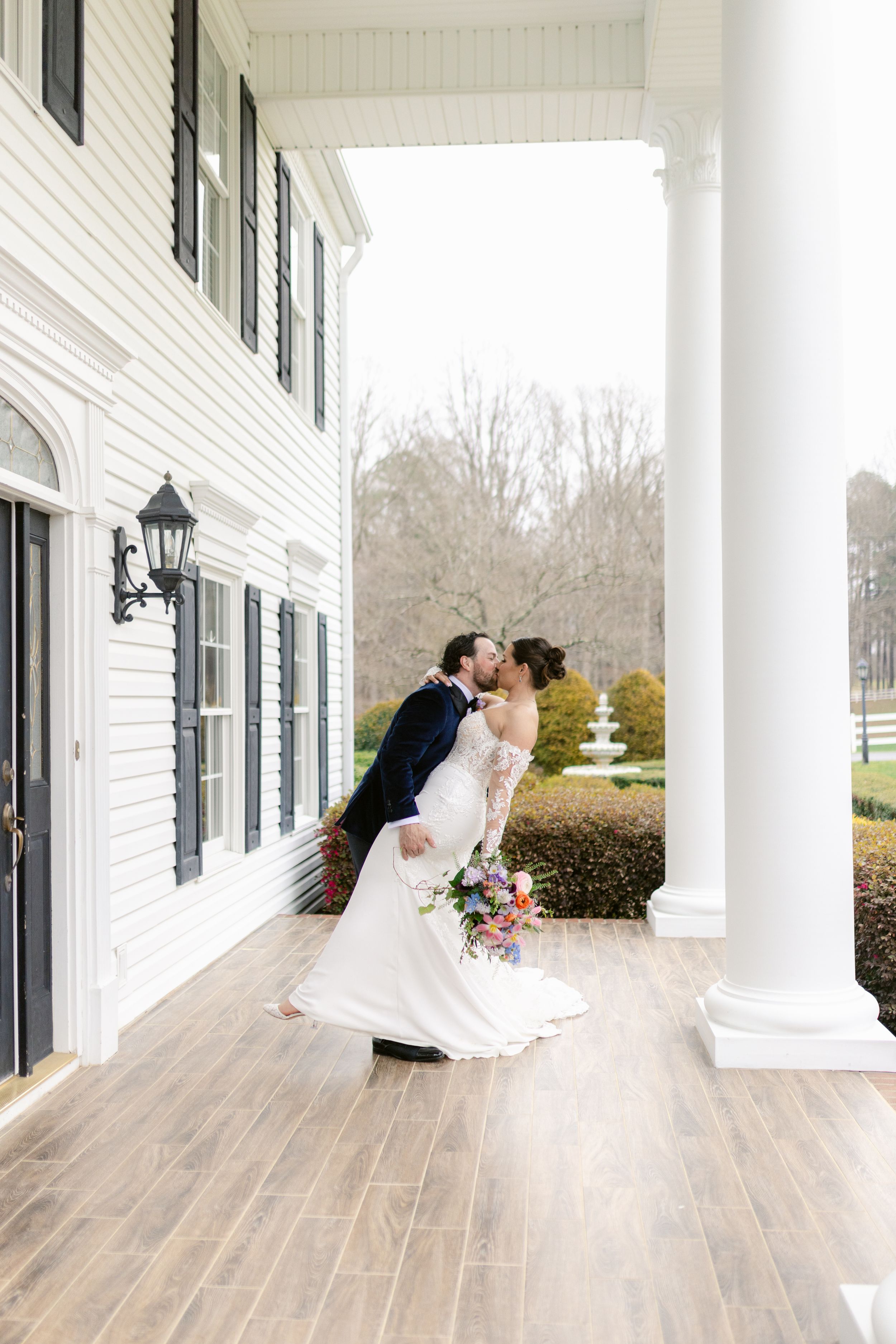 Romantic couple shares tender moments on a white columned porch with wooden floors during their wedding celebration.