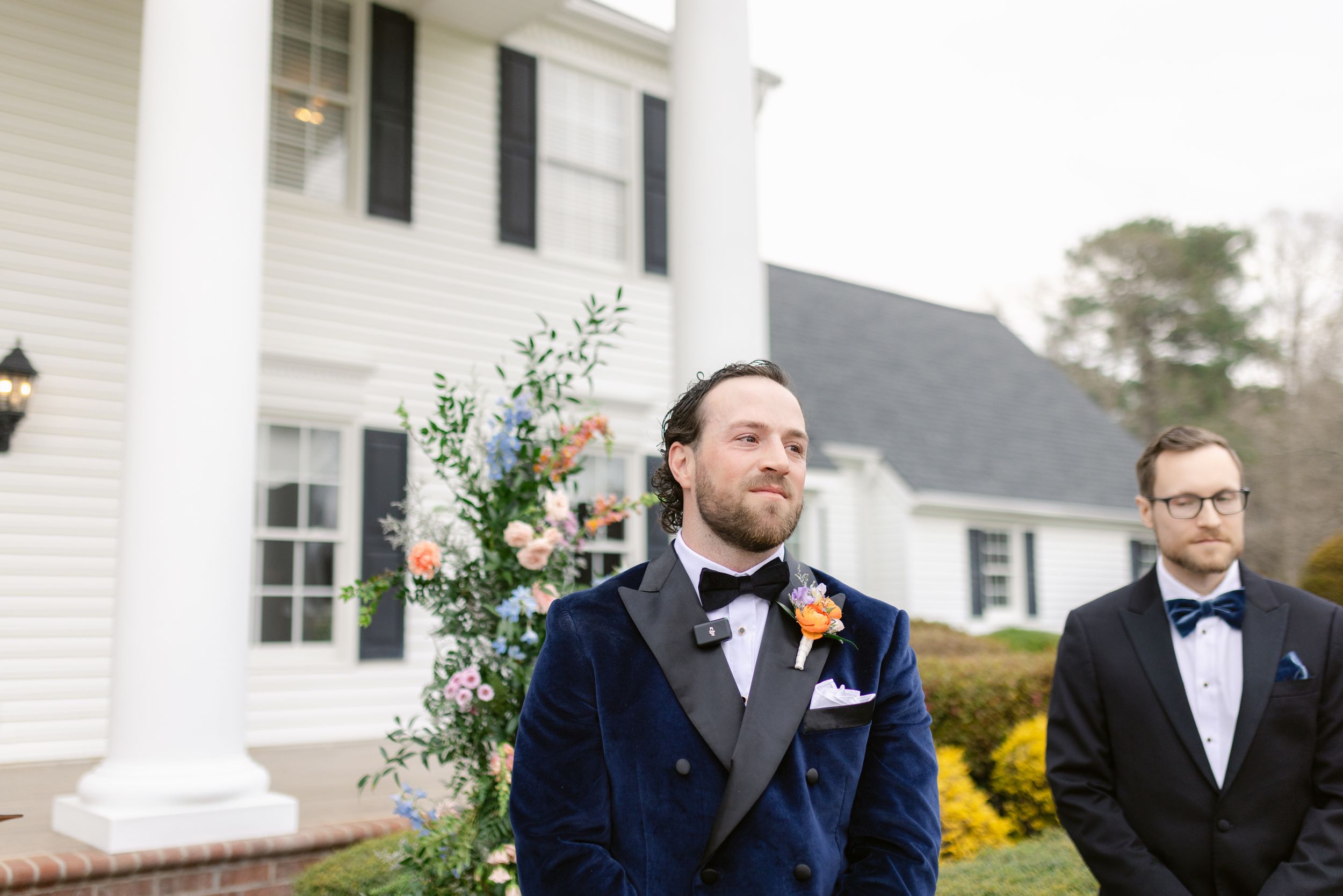 Two people in tuxedos stand outside a white house during a formal event with floral decorations.