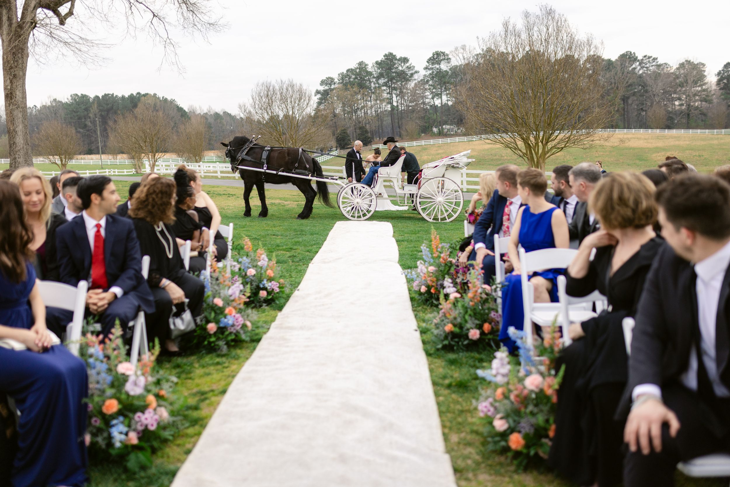 Wedding guests gather outdoors in a scenic countryside setting with trees and rolling hills in the background.