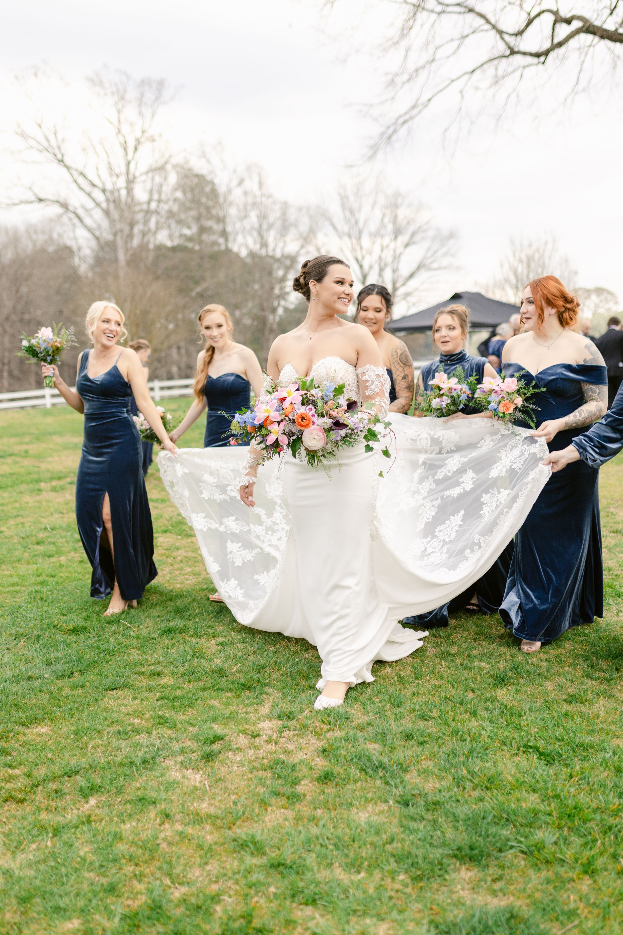 Bridesmaids in navy dresses help bride with flowing white dress twirl dramatically on grassy hillside.