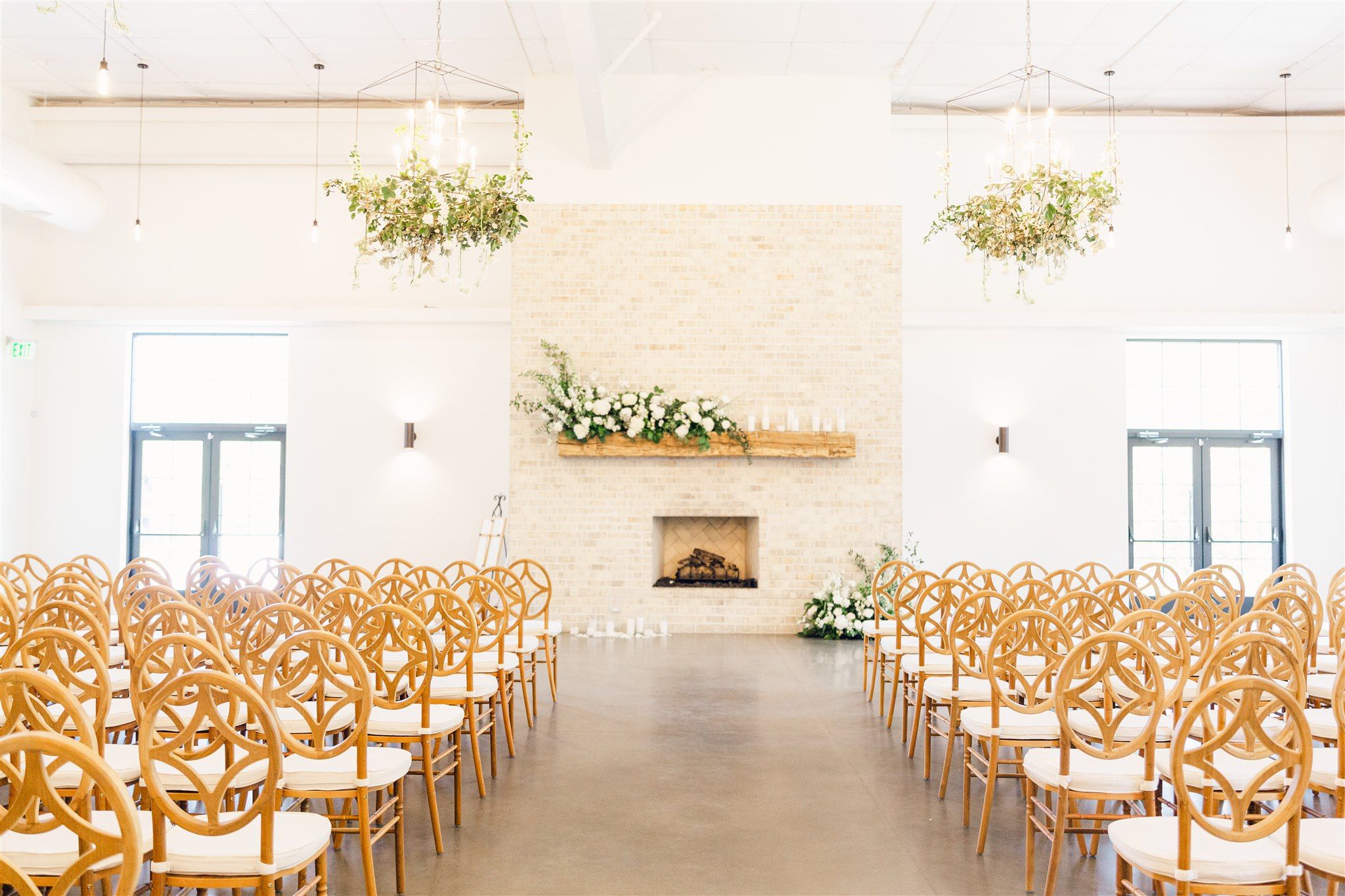 A bright white wedding ceremony space with rows of wooden cross-back chairs and elegant floral arrangements.