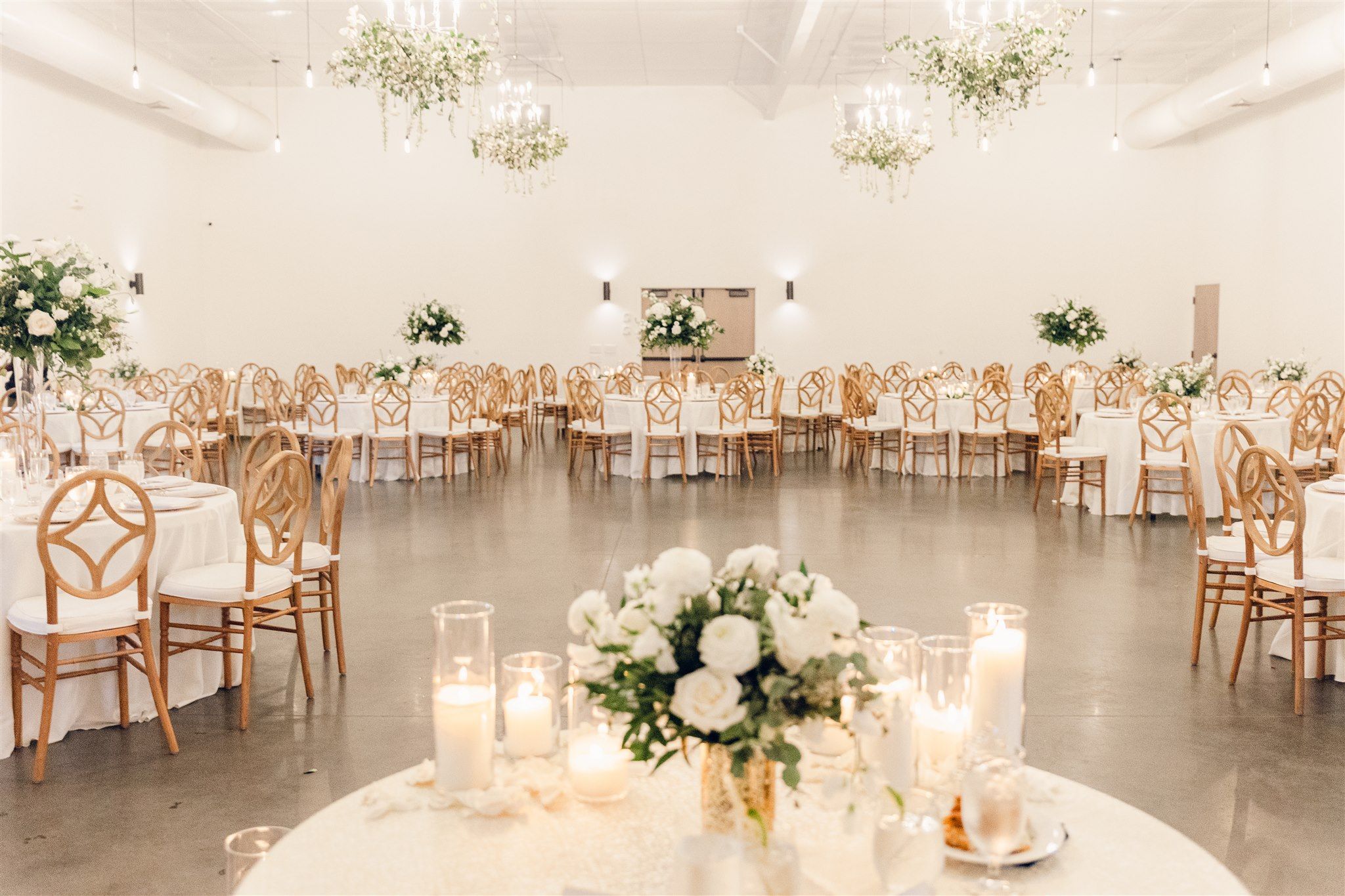 Elegant white wedding reception venue with wooden chairs, floral arrangements and glowing candlelight creating a romantic atmosphere.
