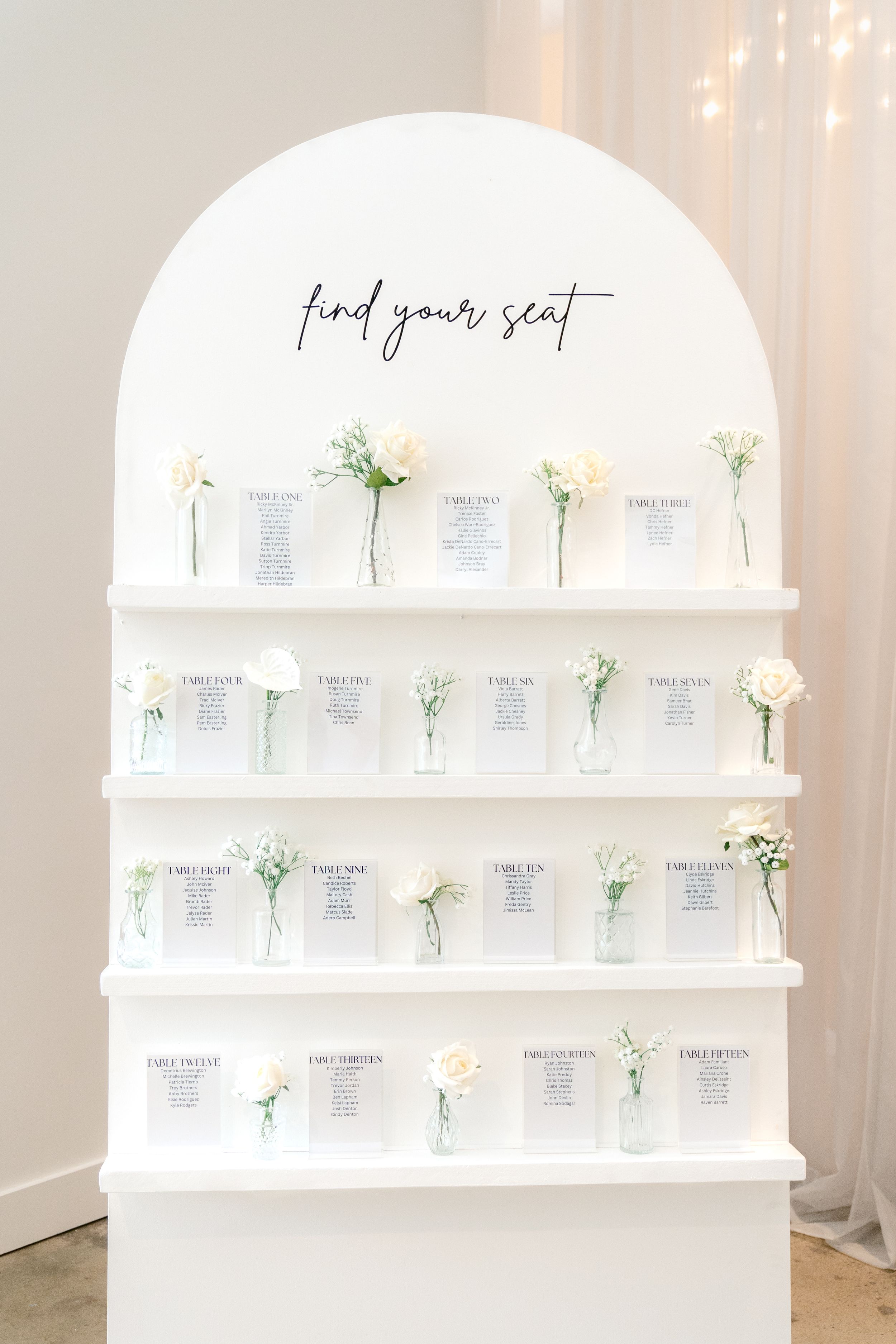 White arched seating chart display with delicate floral accents and paper cards arranged on elegant curved shelves.