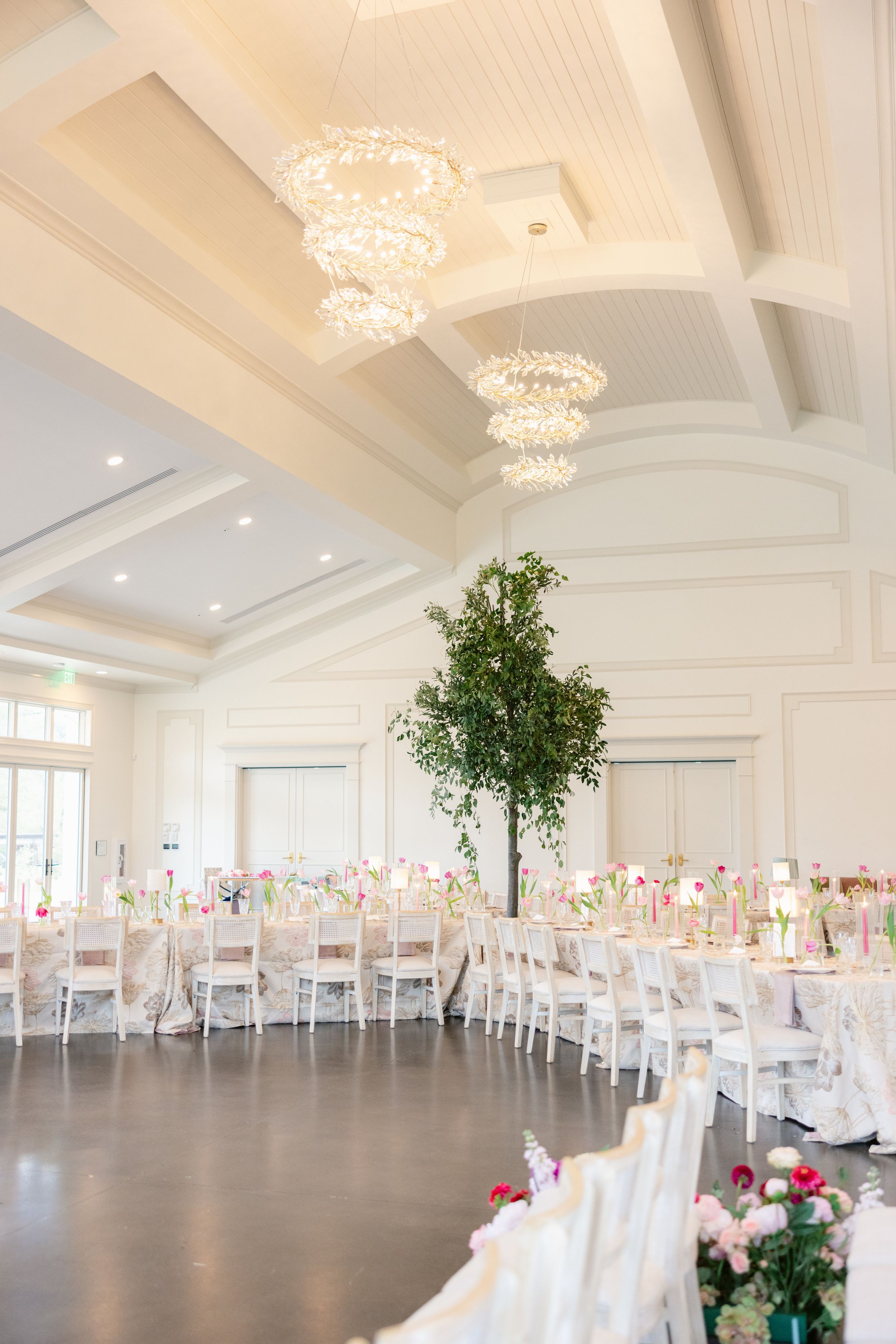 An elegant white wedding reception hall with crystal chandeliers and rows of tables set for a celebration.