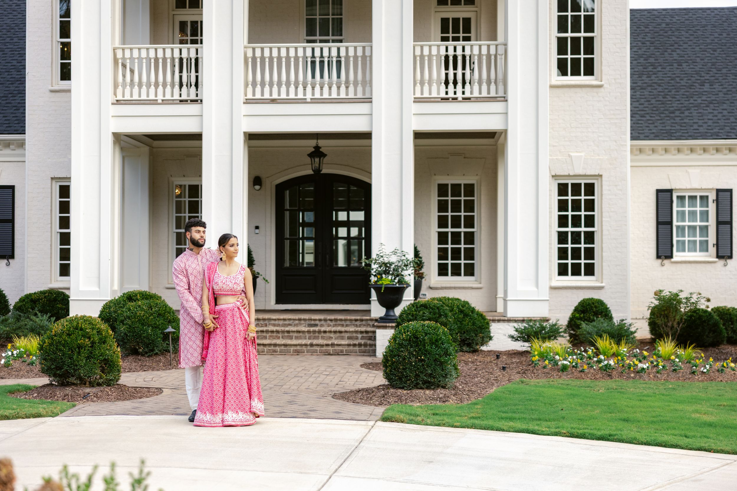 White colonial style mansion with columns and balconies stands behind manicured lawn and landscaping.