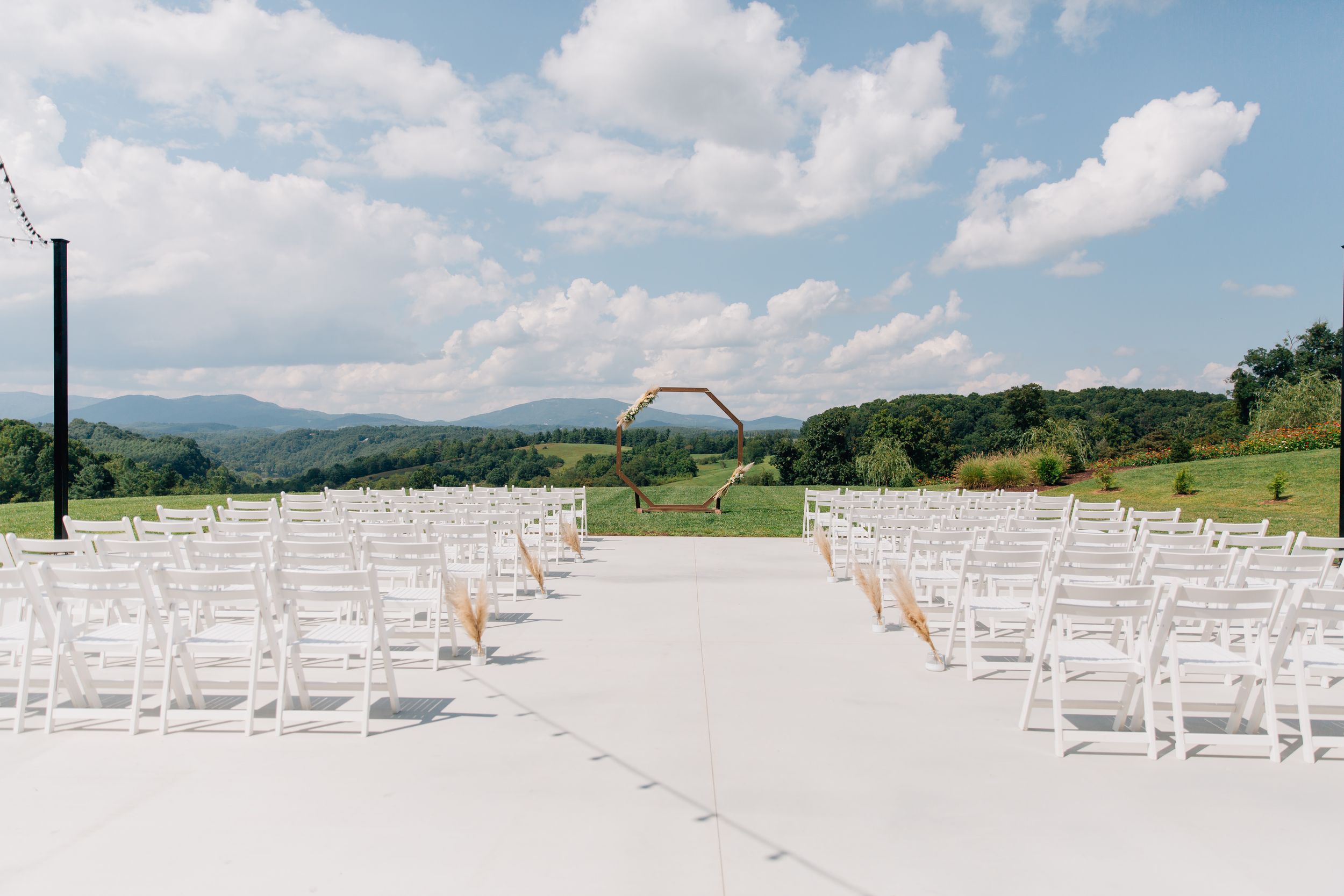 White wooden chairs arranged in rows for an outdoor wedding ceremony with mountains and cloudy sky in the background.
