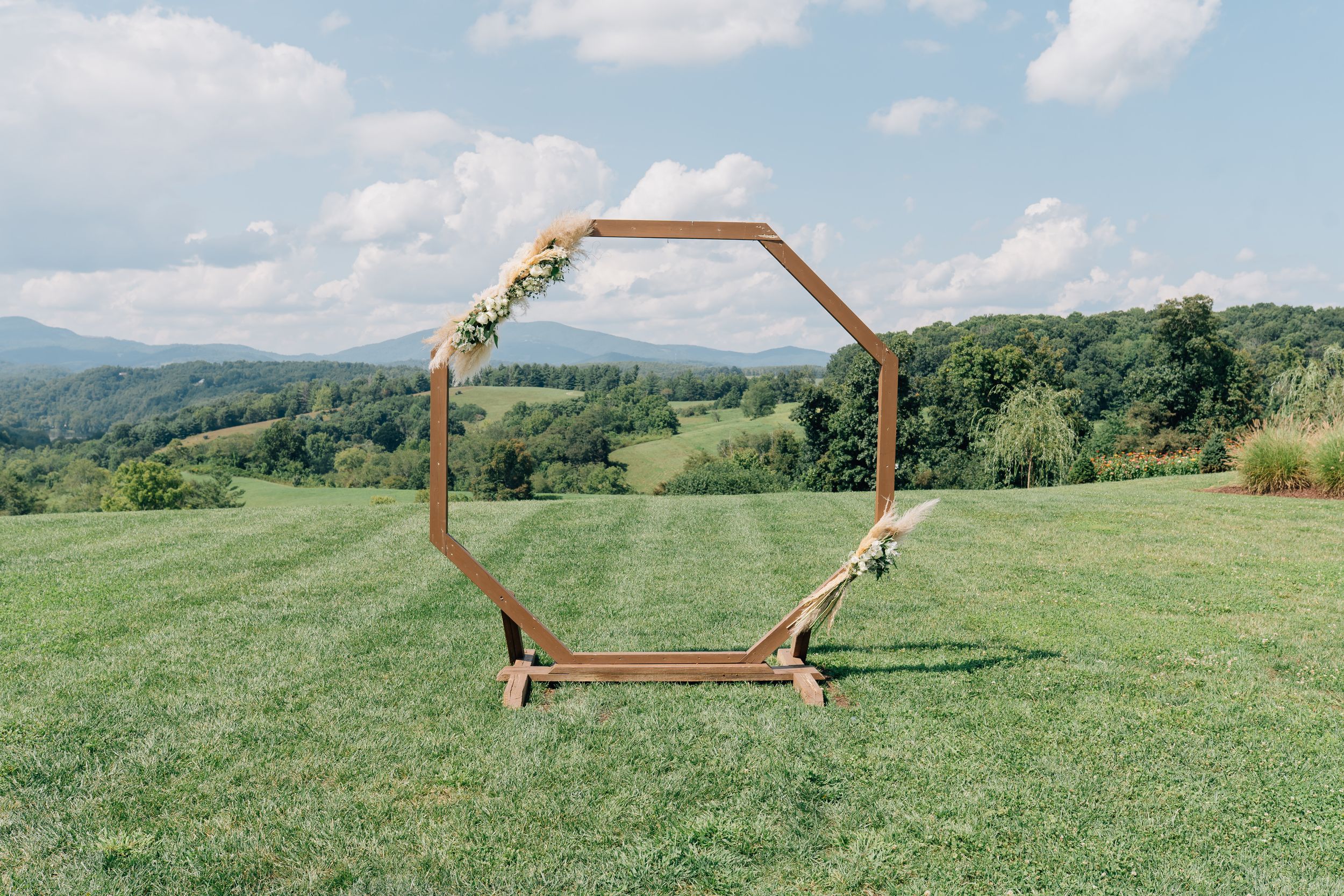 A wooden arch decorated with flowers stands in a green meadow with rolling hills and mountains in the background.