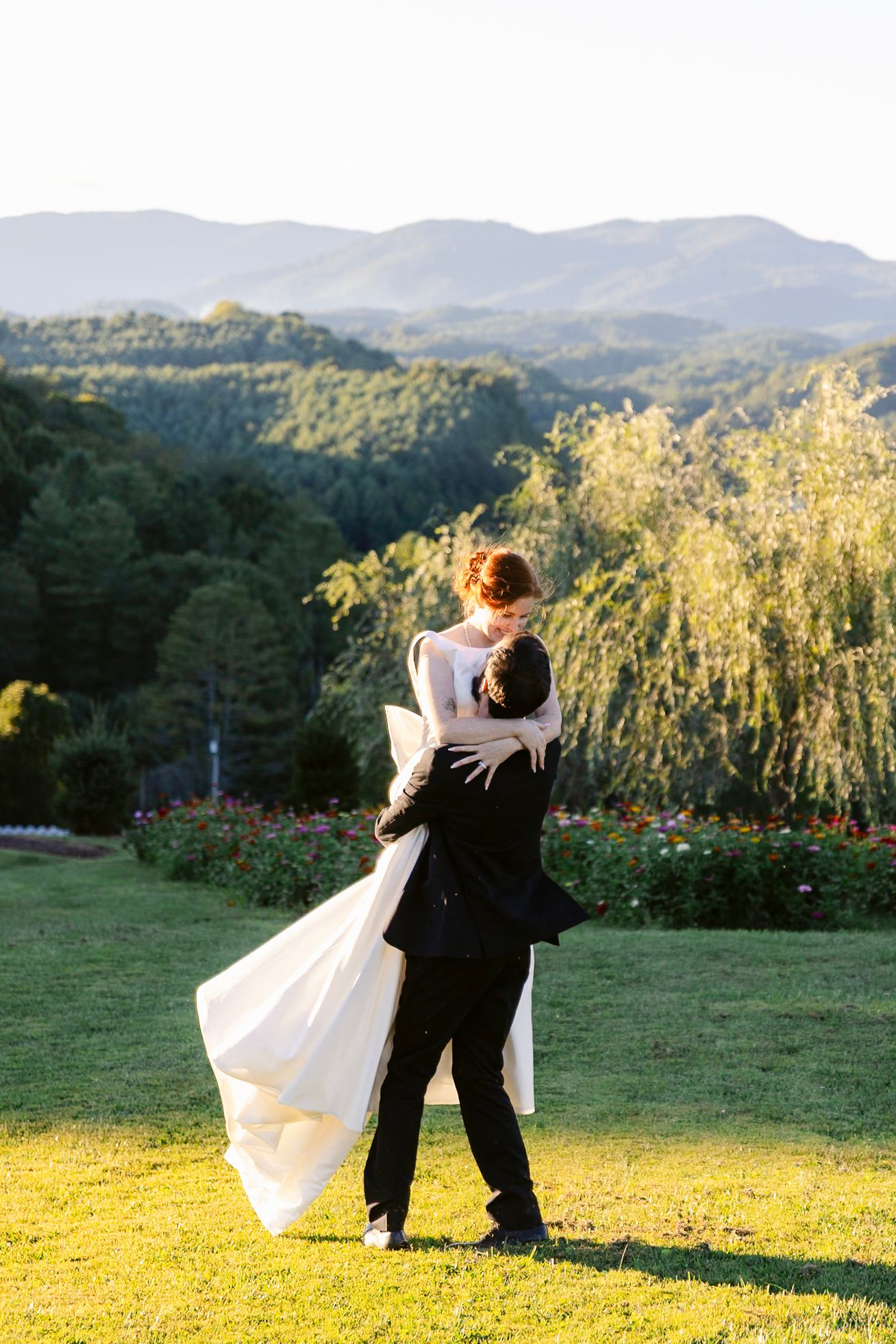 A couple in wedding attire dances together on a hilltop lawn with mountains and golden sunlight in the background.