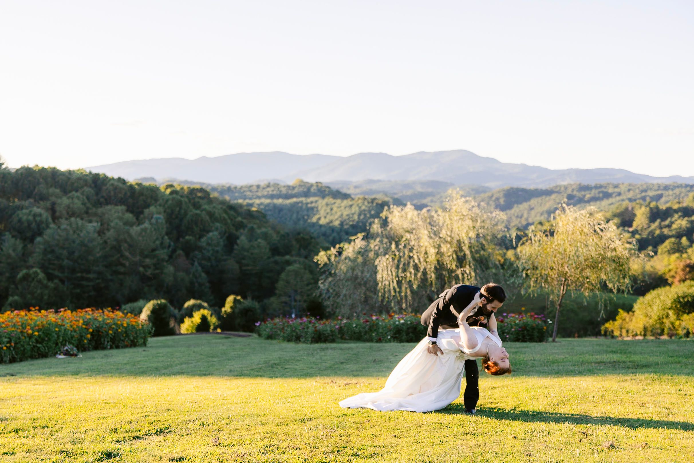 Couple in wedding attire embraces on a scenic hilltop meadow at sunset with mountains in the background.