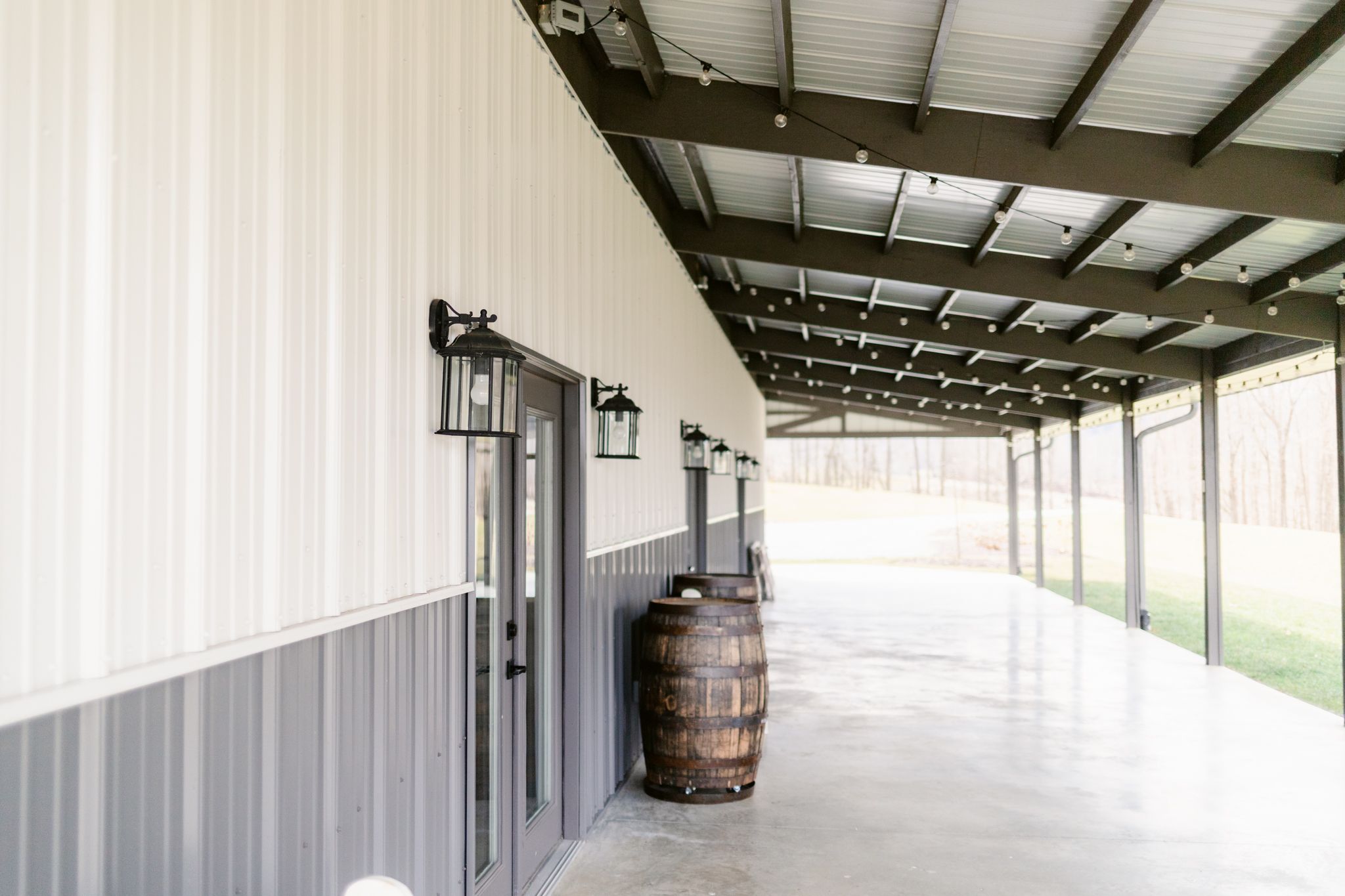 Long covered walkway with white walls and wooden barrels along a rustic building exterior.