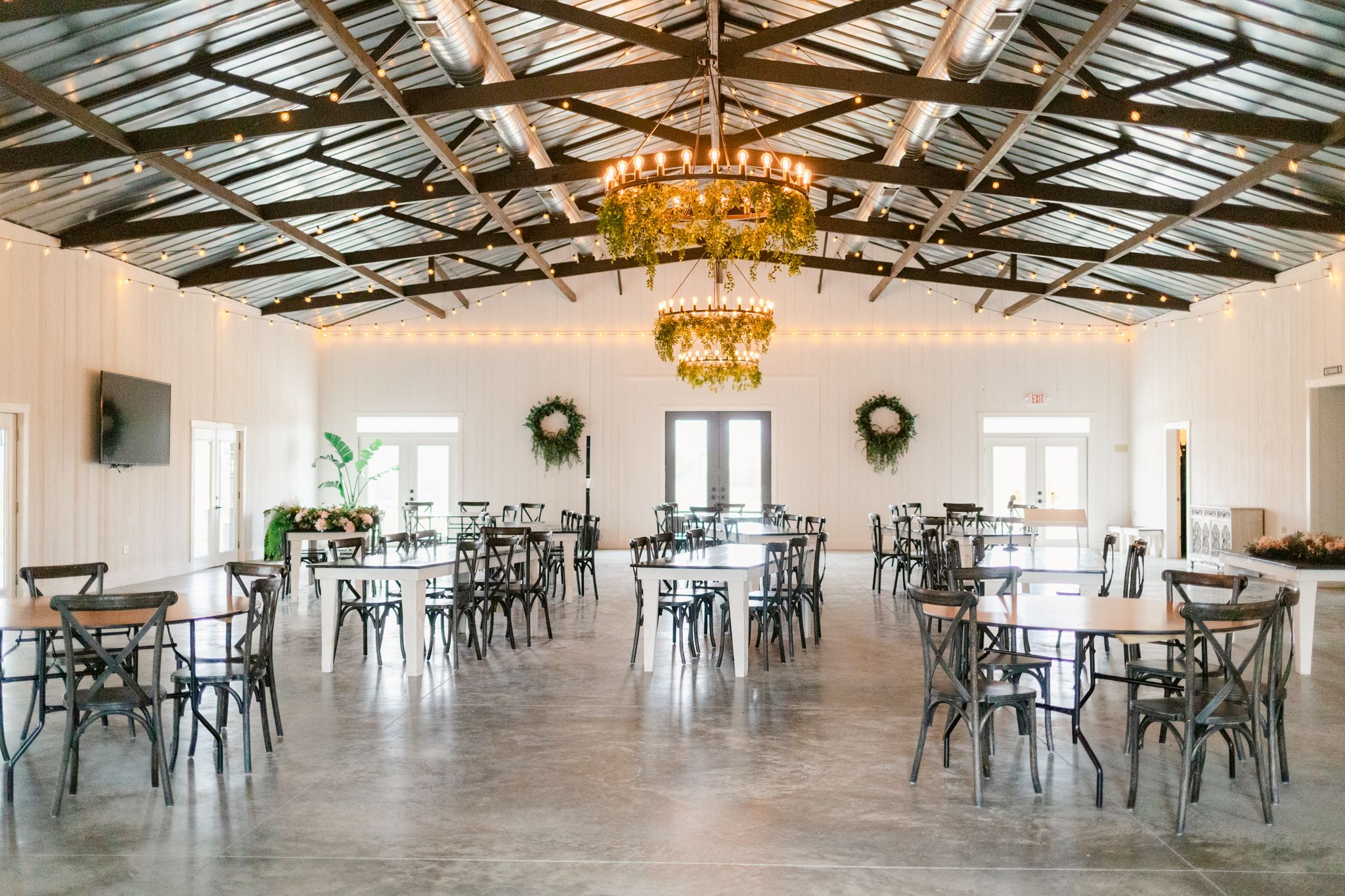 Large white reception hall with exposed wooden beams, chandeliers, and scattered tables and chairs for events.